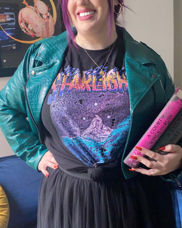 person standing wear the City of Starlight Vintage Location Tee from A Court of Thorns and Roses Inside Green Jacket with a skirt while holding the books in one hand and on their hip with the other