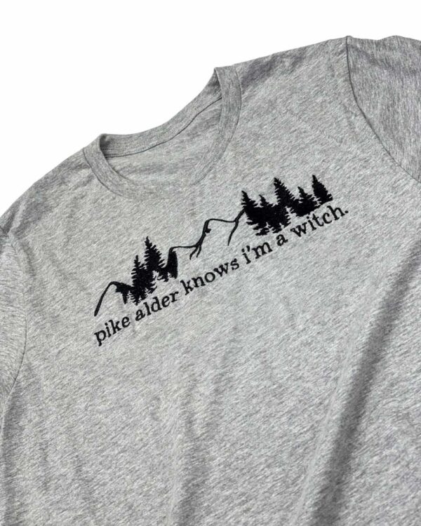 Close up of gray shirt with embroidered design reading "pike alder knows i'm a witch." inspired by Rachel Griffin's Wild is the Witch.